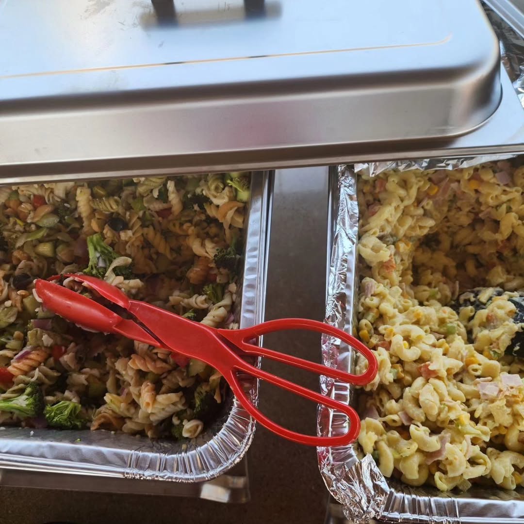 Two foil trays of pasta salad set up in a chafing rack with serving tongs